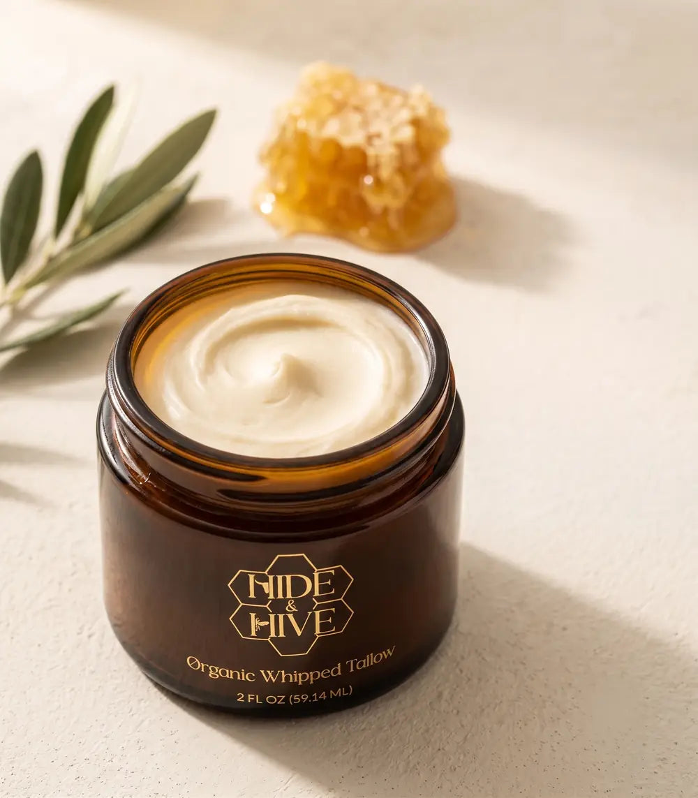 An open jar of Hide & Hive Organic Whipped Tallow in amber glass, its pale whipped surface swirled and catching morning light, with a sprig of olive and a piece of raw honeycomb resting behind it.